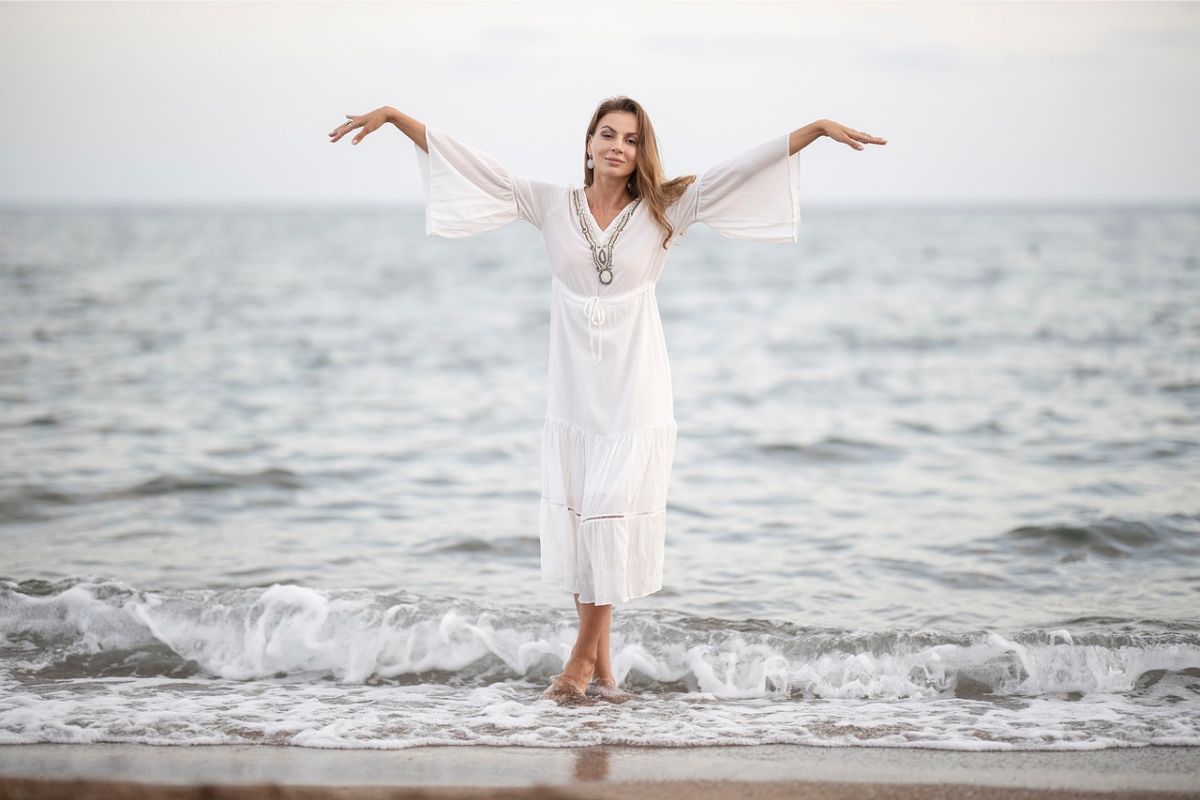 woman wearing a flowy white dress at the beach with her hands in the air